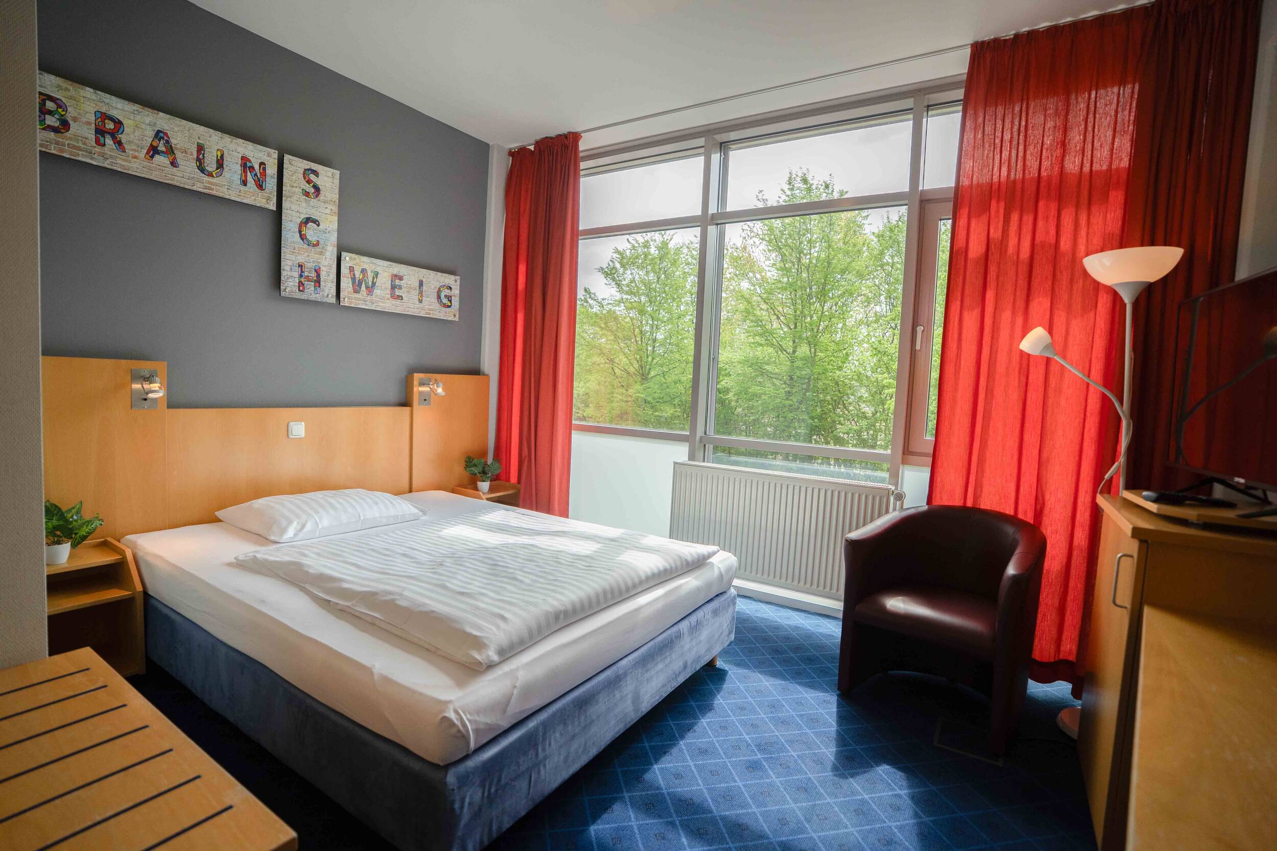 Helles Hotelzimmer mit Doppelbett, rotem Vorhang, Sessel, Schreibtisch und großem Fenster mit Blick ins Grüne.
