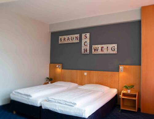 Großes Doppelbett aus warmen Holz vor einer modernen grauen Wand. Über dem Bett hängt ein Schriftzug im Industrial-Design.