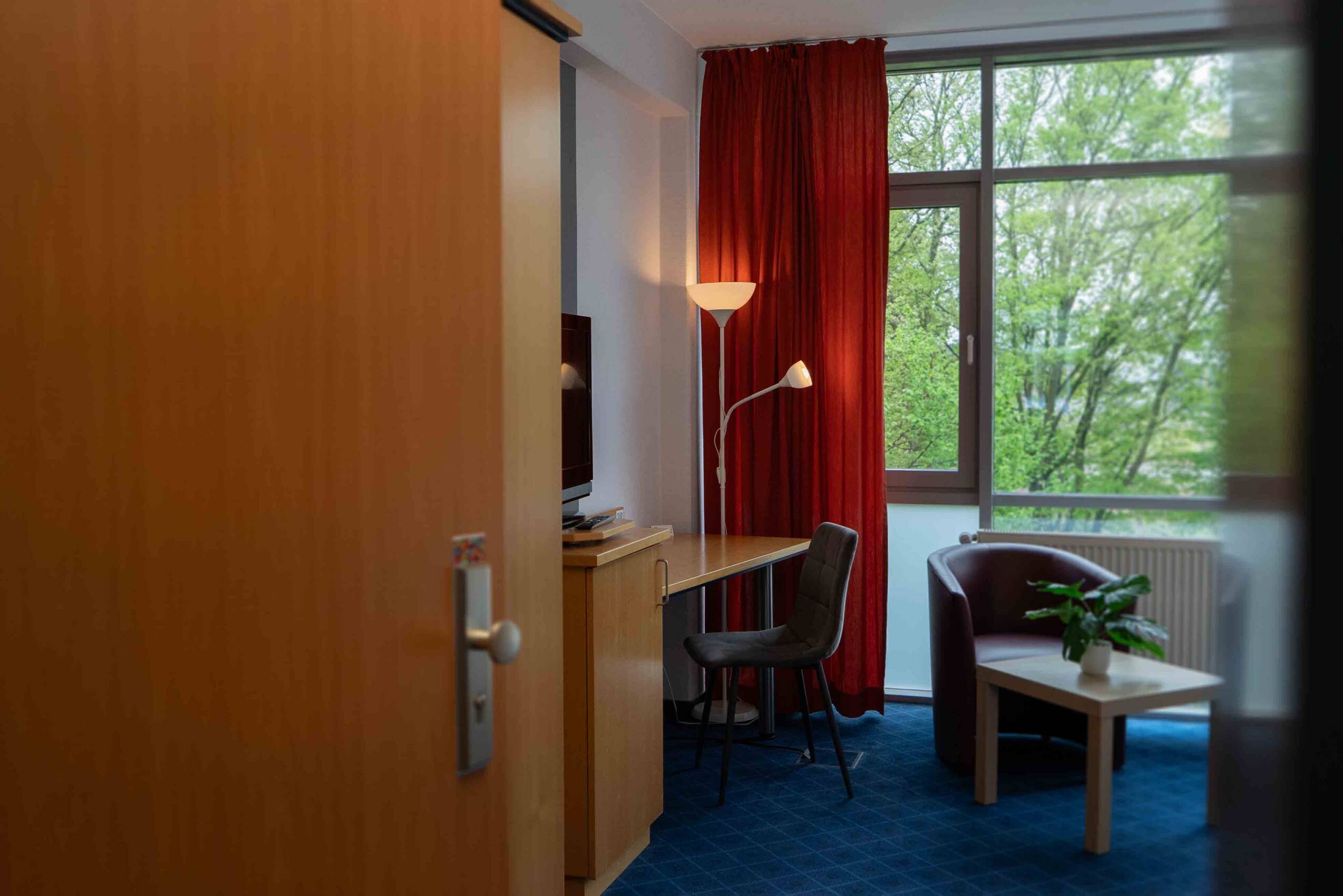 Hotelzimmer mit warmen Holzmöbeln, Flachbild-TV, großzügigen Schreibtisch und gemütlicher Sitzecke vor einem bodentiefen Fenster.