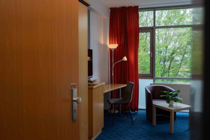 Hotelzimmer mit warmen Holzmöbeln, Flachbild-TV, großzügigen Schreibtisch und gemütlicher Sitzecke vor einem bodentiefen Fenster.