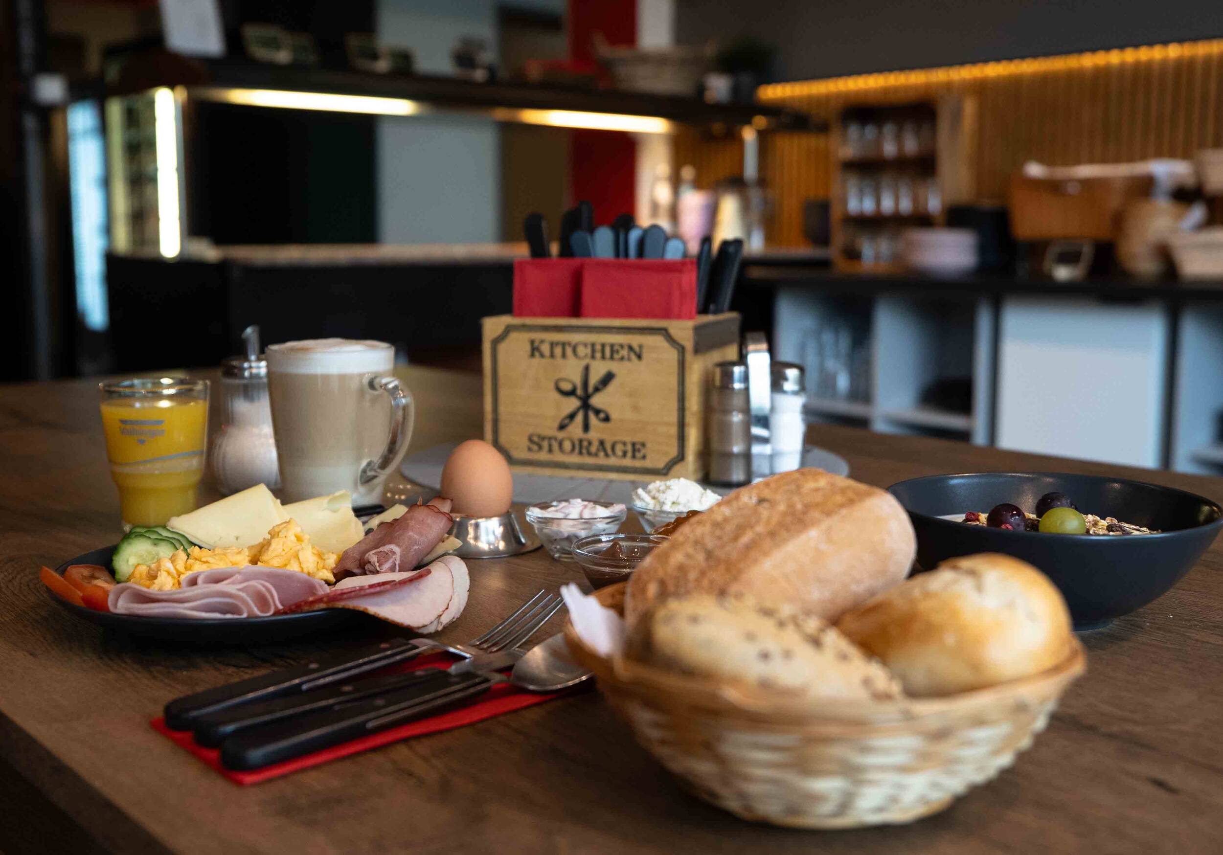 Gedeckter Tisch im Frühstücksraum mit allerlei Aufschnitt, einem Frühstücksei, frischen Getränken und einem Korb voller knuspriger Brötchen.
