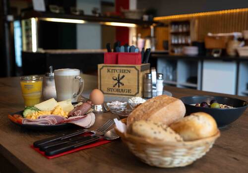 Gedeckter Tisch im Frühstücksraum mit allerlei Aufschnitt, einem Frühstücksei, frischen Getränken und einem Korb voller knuspriger Brötchen.